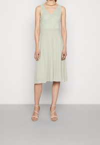 Ljusgrön knälång klänning med en spetsöverdel och en plisserad kjol; har en V-ringning och matchas med beige klackar med remmar.