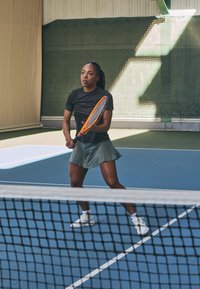 A player in a black shirt and green skirt holds an orange tennis racket, poised to hit a ball on a blue indoor court.