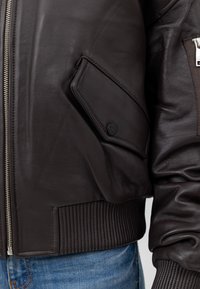 Brązowa skórzana kurtka bombowa z zamkiem błyskawicznym, żebrowanymi mankietami oraz kieszenią na rękawie z klapką i czarnym guzikiem jako akcentem.