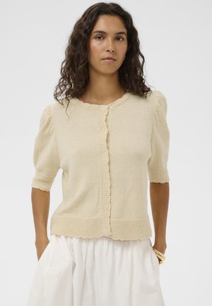 Femme aux cheveux bruns bouclés portant un cardigan beige à manches courtes en tricot et une jupe blanche, debout devant un fond uni.