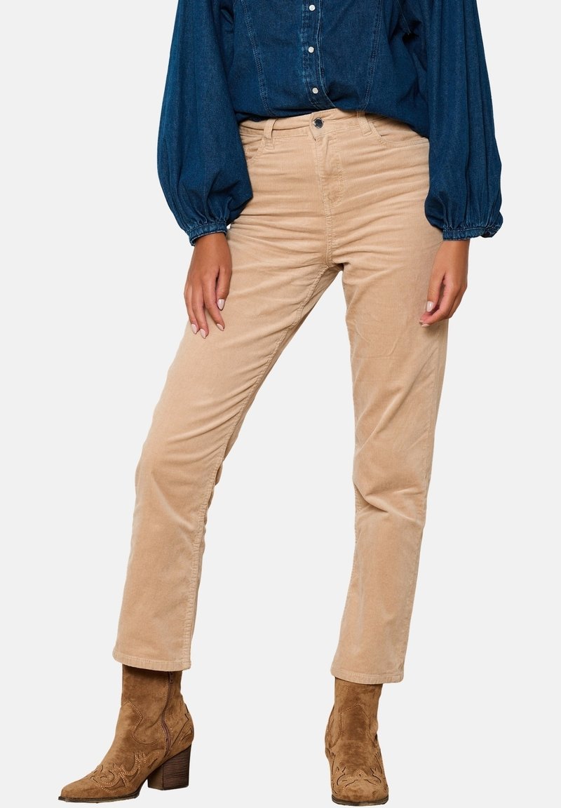 Personne portant un pantalon en velours côtelé beige, des bottines marron en daim à talons et une chemise bleu foncé à manches longues bouffantes sur un fond uni.
