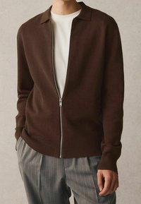 Cardigan en tricot marron avec une fermeture éclair à l'avant, des poignets et un ourlet côtelés, et un col plat, associé à un pantalon gris à fines rayures.