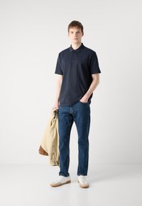 Tricou polo bleumarin cu guler și placket cu trei butoane, asortat cu blugi albaștri și adidași albi; ținând în mână o jachetă bej.