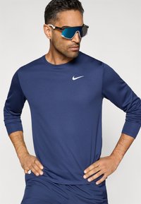 Μακρυμάνικο αθλητικό πουκάμισο σε ναυτικό μπλε χρώμα, κατασκευασμένο από ελαφρύ ύφασμα, με διακριτικό λογότυπο Nike σε λευκό. Φοριέται με ανακλαστικά μπλε γυαλιά ηλίου.