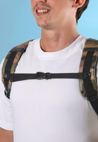 Spalline di uno zaino con motivo camouflage, dotate di una fascia sternale nera e una fibbia, indossate sopra una maglietta bianca su uno sfondo blu.