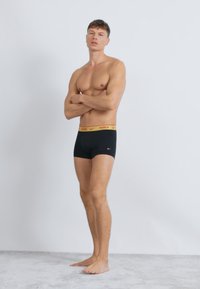 Čierne elastické bavlnené boxerky s pásom s logom Nike v žltej farbe. Model stojí so založenými rukami a predvádza priliehavý strih.