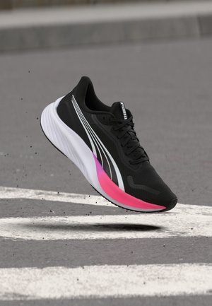 Chaussure de course noire avec des accents blancs et roses sur la semelle, logo Puma blanc sur le côté, montrée en vol au-dessus d'une surface asphaltée.