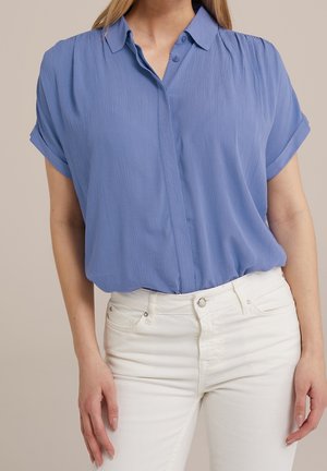 Vrouw draagt een lichtblauwe blouse met korte mouwen, ingestopt in een witte, hooggetailleerde spijkerbroek tegen een neutrale achtergrond.