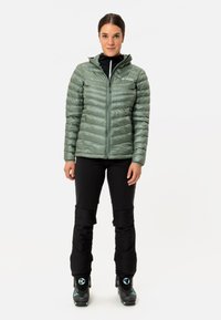 Groene pufferjas met horizontale stiksels, ritssluiting en capuchon. Draag het over zwarte skibroeken en laarzen, wat een functionele outdoorlook laat zien.