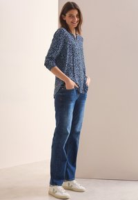 Blaue gemusterte Bluse mit Knopfleiste und Dreiviertelärmeln, kombiniert mit dunkelblauen geraden Jeans und weißen Turnschuhen.