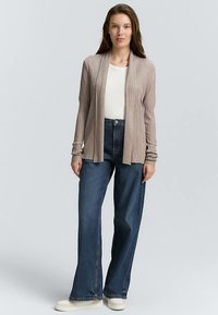 Lang ribstrikket cardigan i lys taupe, båret over en hvid top, kombineret med mørkeblå viddebukser og lyse sneakers.