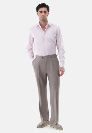 Mann steht, trägt ein hellrosa kariertes Hemd, hoch taillierte graue plissierte Hose und beige Slipper vor weißem Hintergrund.