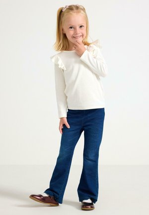 Weißes Langarmoberteil mit Rüschen an den Schultern, kombiniert mit dunkelblauen Schlagjeans und braunen Schuhen. Einfaches, lässiges Outfit für Kinder.