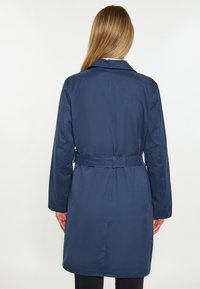 Granatowy płaszcz trencz z paskiem, z klasycznym kołnierzem, długimi rękawami i gładkim, wodoodpornym materiałem. Widok z tyłu ukazuje strzałkowy design.