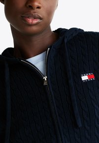 Marine blauwe zip-up hoodie in structuur kabelbreisel met een zilveren rits en een klein rood, wit en blauw logo patch op de borst.