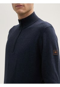 Navyblauer Halb-Reißverschluss-Pullover aus strukturiertem Stoff, mit hohem Kragen und einem braunen Logo-Patch am linken Ärmel.