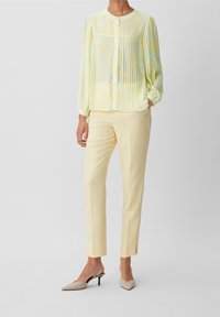 Blusa a pieghe giallo chiaro con bottoni, abbinata a pantaloni gialli attillati. Le scarpe sono beige con punta affusolata e tacco basso. Sfondo semplice.