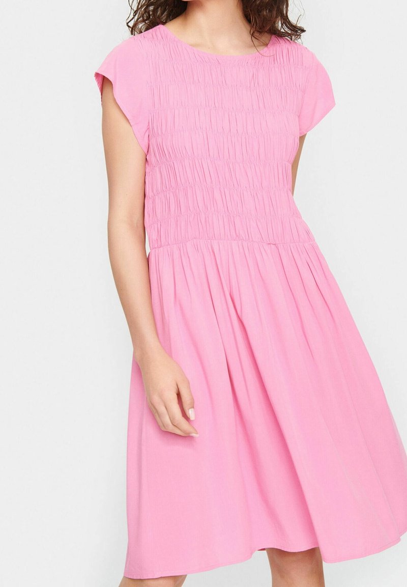 Femme portant une robe courte rose à manches courtes, longueur aux genoux, avec un corsage smocké et une jupe froncée, debout devant un fond uni.