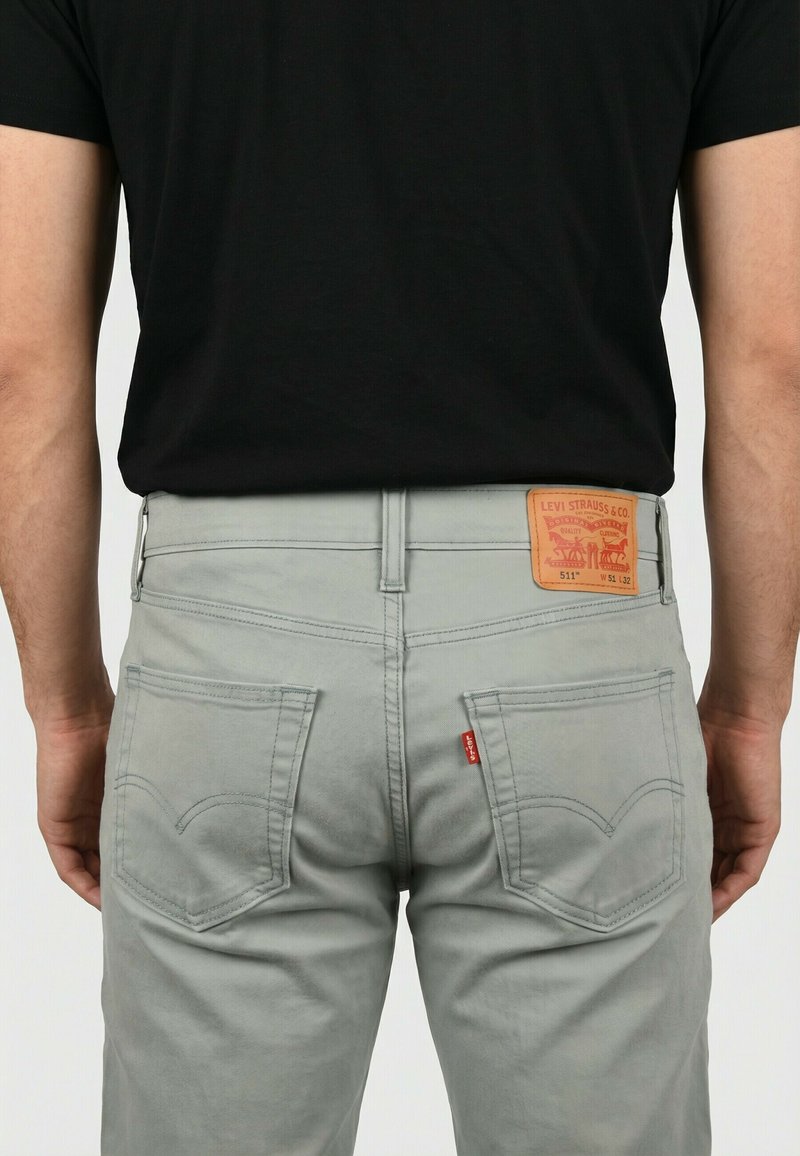 Hombre con vaqueros Levi's 511 gris claro y camiseta negra, visto desde la cintura hacia abajo por detrás, de pie frente a un fondo liso.