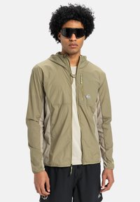 Lettet olivengrøn jakke med hætte, lynlåslomme foran og beige detaljer. Båret over en hvid skjorte. Sorte solbriller fuldender looket.