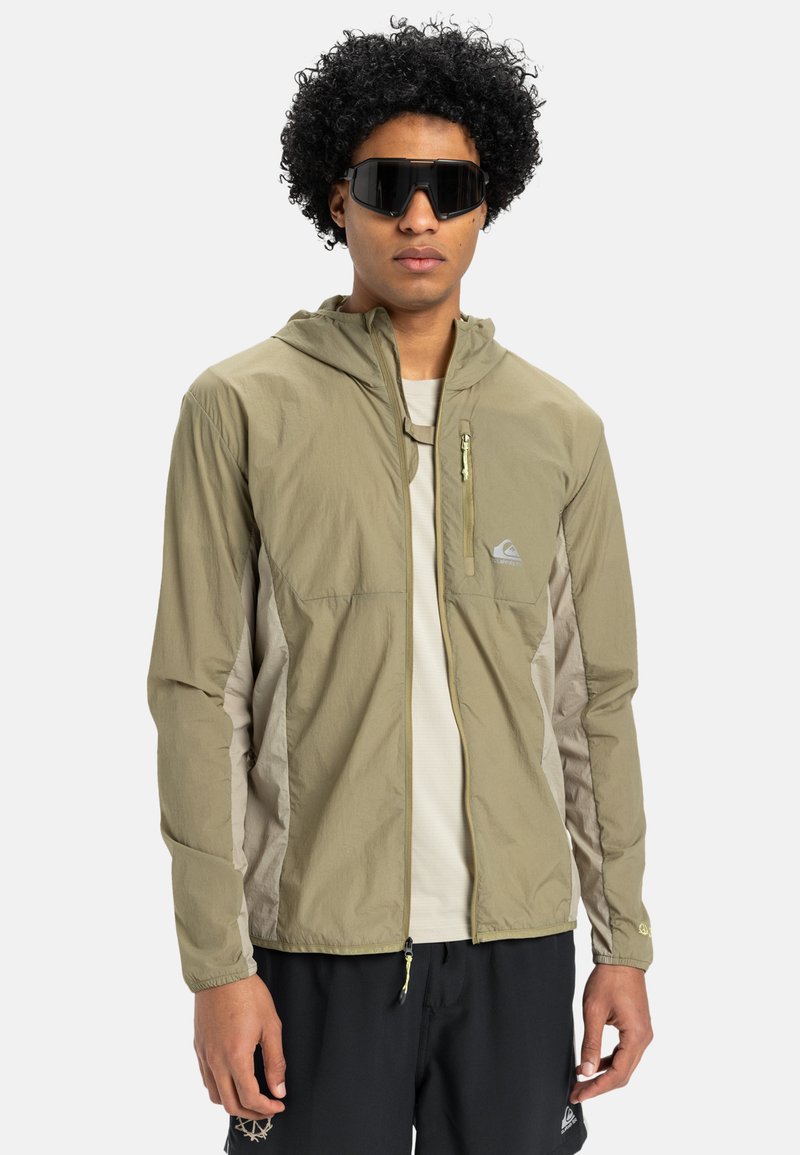 Lettet olivengrøn jakke med hætte, lynlåslomme foran og beige detaljer. Båret over en hvid skjorte. Sorte solbriller fuldender looket.