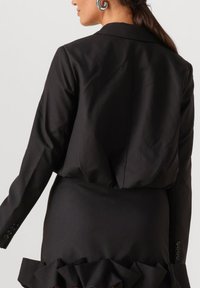 Zwarte op maat gemaakte jas met een gestructureerd ontwerp, voorzien van een enkele split en gecombineerd met een nauwsluitende rok versierd met ruches aan de zoom.