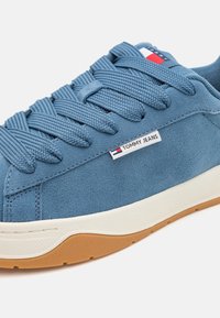 Blå semsket sneakers med teksturerte, vevde snørebånd, hvit gummisåle og brune detaljer. Har en liten "TOMMY JEANS" etikett på siden.