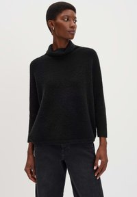 Schwarzer Cowl-Neck-Pullover aus strukturiertem Strick, mit langen Ärmeln und lockerer Passform. Kombiniert mit dunklen Jeans.