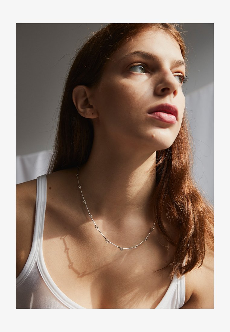 Silverkedja med avlånga länkar och cirkulära detaljer, bärs med en enkel vit linne mot en mjukt belyst bakgrund.