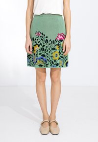 Femme portant une jupe florale verte avec de grandes fleurs colorées, associée à des chaussures Mary Jane beiges à boucle, se tenant devant un fond blanc.