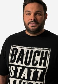 Černé bavlněné tričko s velkým bílým roztrhaným potiskem textu, který říká "BAUCH STATT NICHT." Krátké rukávy a kulatý výstřih.