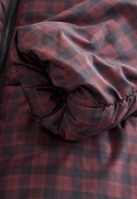 Doudoune en textile avec motif à carreaux rouge foncé et bleu marine, présentant une texture douce et froissée avec un détail de tissu froncé près de la fermeture éclair.