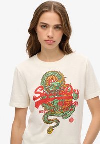 Camiseta de algodón crema con un gráfico colorido de dragón en verde, rojo y amarillo, con elementos de texto en negrita impresos en la parte frontal.