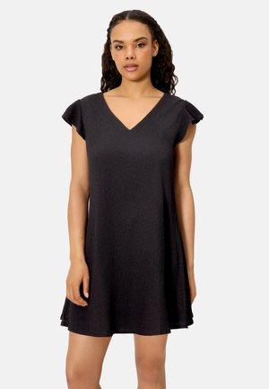 Mujer con un vestido corto negro texturizado con mangas con vuelo y escote en V, de pie contra un fondo claro y liso.