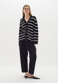 Randig svart och vit cardigan med V-ringning och fem stora knappar, tillsammans med lösa svarta byxor och svarta spetsiga plattor.