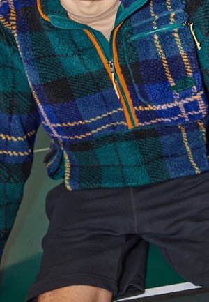 Persona con una chaqueta polar a cuadros verdes y azules, con cremallera naranja y pantalones cortos oscuros, sentada sobre una superficie verde.