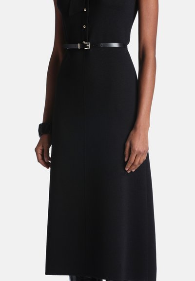 Femme portant une robe noire sans manches avec des boutons et une fine ceinture noire à la taille, debout devant un fond uni.