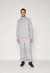 Gray tracksuit with zip jacket and elastic cuffs, featuring white accents and logo. The pants have an elastic waistband, paired with black sneakers.