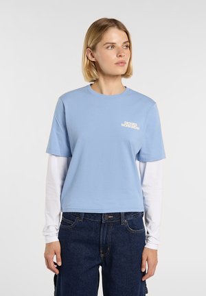 Jasnoniebieska bawełniana koszulka z okrągłym dekoltem i krótkimi rękawami, z białym napisem "Dickies Workwear" na piersi, noszona na białej koszulce z długim rękawem.