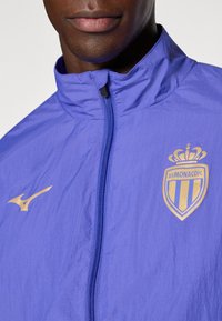 Violeta gaisā jaka ar augstu apkakli, ar rāvējslēdzēju, zelta akcentiem, Mizuno logo un AS Monaco FC emblēmu. Gluda tekstūra un izturīgs dizains.