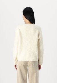 Krämfärgad, mjuktexturerad tröja med långa ärmar, rund halsringning, ribbat fåll och avslappnad passform. Bärs över kordflorsbyxor.