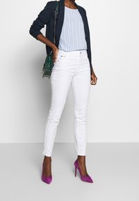 Jeans ajustados blancos, camiseta de rayas en azul claro, blazer azul marino y tacones morados. Sostiene un bolso verde con cadena.