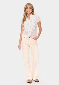 White sheer blouse with ruffled front detail, lightweight fabric, paired with light beige wide-leg trousers and black strappy sandals.