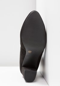 Chaussure en daim noir à bout pointu avec talon carré, semelle indiquant la taille 37, positionnée debout contre un coin blanc.
