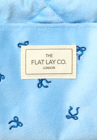 Tela de terciopelo azul claro con pequeños lazos bordados en azul. La etiqueta dice "THE FLAT LAY CO. LONDON" en negro sobre un fondo blanco.