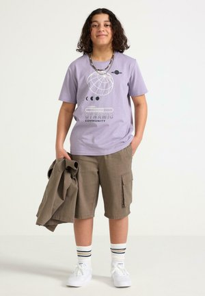 Smilende person med krøllet hår iført en lilla t-shirt med grafik, beige cargoshorts, hvide sneakers, stribede sokker og holder en matchende jakke.