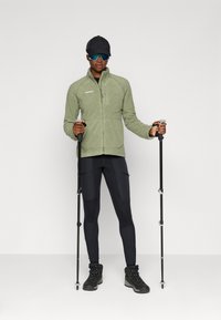 Grüne Fleecejacke mit Reißverschluss, schwarze Leggings und schwarze Wanderschuhe. Hält Trekkingstöcke und trägt Sonnenbrillen und eine schwarze Mütze.