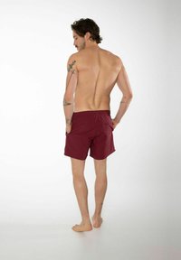 Shorts de bain maroons avec un design uni, une taille élastique et des poches latérales, vus de l'arrière. Texture lisse et longueur au-dessus du genou.