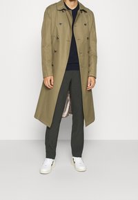 Mann in khakifarbenem doppelreihigem Trenchcoat über einem schwarzen Pullover, dunkelt Grauen Hosen und weißen Sneakers vor einem schlichten weißen Hintergrund.