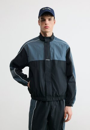 Männliches Modell in einem zweifarbigen schwarz-grauen Calvin Klein Trainingsanzug mit weißen Streifen, trägt eine marineblaue Kappe und steht vor einem schlichten weißen Hintergrund.